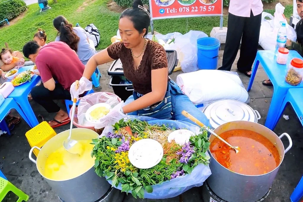 A street food vendor in Siem Reap ladles curry beside a platter of fresh herbs for nom banh chok rice noodles
