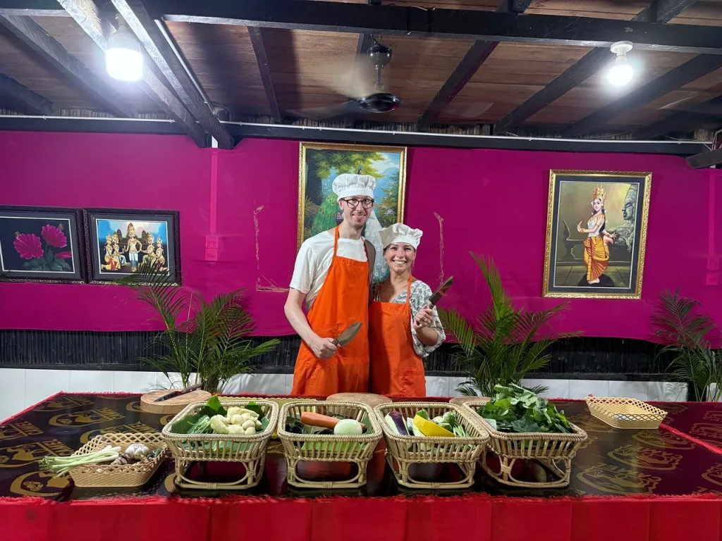 Two guests in chef's hats and orange aprons at the cooking class table, with baskets of fresh ingredients ready to prepare